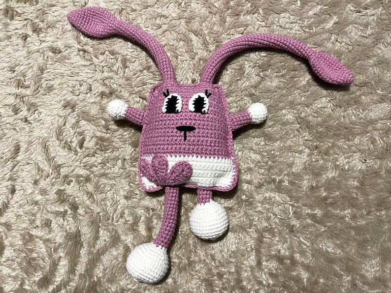 May include: A pink crocheted bunny toy with white accents and black eyes. The bunny has long ears and a heart-shaped detail on its chest.