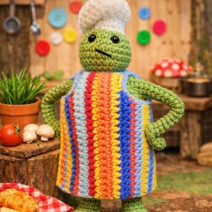 May include: A crocheted frog chef doll wearing a white chef's hat and a colorful striped apron. The frog is standing in a kitchen setting with a tomato, mushrooms, and a croissant on a wooden surface.