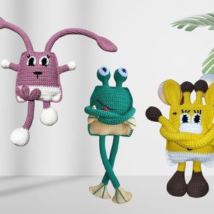 May include: Three handmade crochet toys: a purple bunny, a green frog, and a yellow giraffe. The bunny has long ears and dangling legs. The frog has large eyes and crossed arms. The giraffe has brown spots and a white belly. All have white accents.