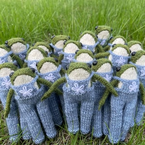 May include: A group of knitted frog dolls with green heads, cream-colored faces, and blue overalls. Each frog has black eyes and green arms. The overalls are decorated with a white flower. The frogs are arranged on a bed of green grass.