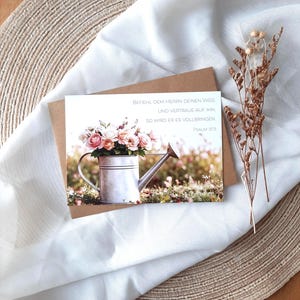 Könnte beinhalten: Eine Grußkarte mit einem Aquarellbild einer Gießkanne, die mit rosa und pfirsichfarbenen Rosen überquillt. Die Karte liegt auf einem weißen Tuch und einer gewebten Matte. Die Karte hat deutschen Text.