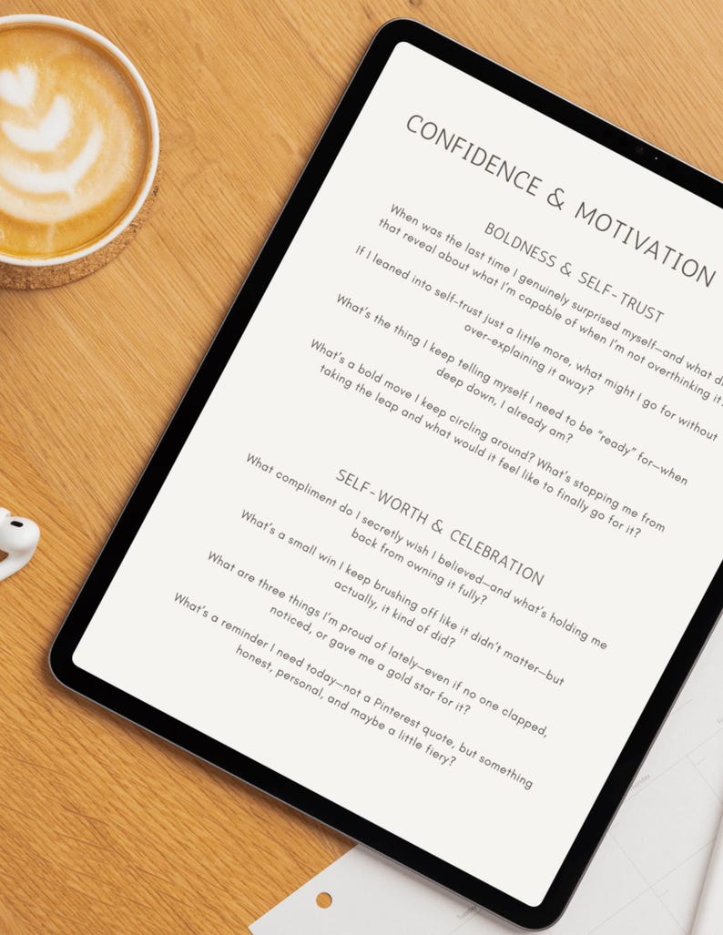 Op de afbeelding: Een digitale tablet met een motivatie werkblad getiteld "Confidence & Motivation". Het werkblad bevat vragen over zelfvertrouwen en zelfwaardering. Een kopje koffie en oortelefoons zijn zichtbaar op een houten oppervlak.