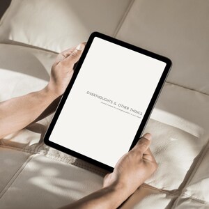 Op de afbeelding: Een zwarte tablet met de tekst "OVERTHOUGHTS & OTHER THINGS". De tablet wordt door een persoon vastgehouden. De achtergrond is een lichtgekleurd, getextureerd oppervlak.