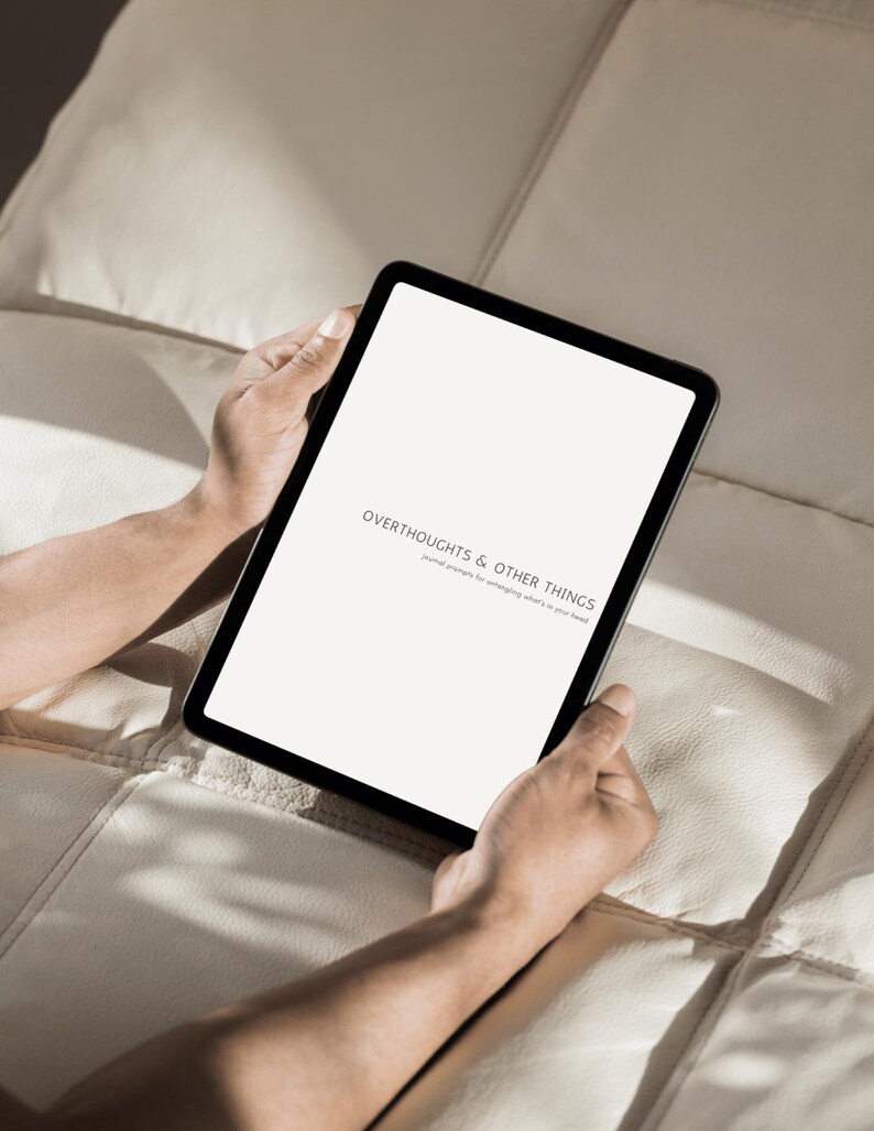 Op de afbeelding: Een zwarte tablet met de tekst "OVERTHOUGHTS & OTHER THINGS". De tablet wordt door een persoon vastgehouden. De achtergrond is een lichtgekleurd, getextureerd oppervlak.