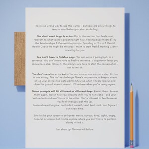 Op de afbeelding: Een dagboek met tekst en een houten basis, vergezeld van een gele en oranje potlood. De pagina's van het dagboek staan vol tekst met richtlijnen voor zelfreflectie en journaling. De achtergrond is lichtblauw.