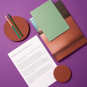 Op de afbeelding: Een stilleven op een paarse ondergrond met een groen notitieboek, een bruin notitieboek, een vel papier met de tekst "MENTAL HEALTH CHECK-INS", twee potloden en twee bruine onderzetters. De potloden zijn groen en oranje.