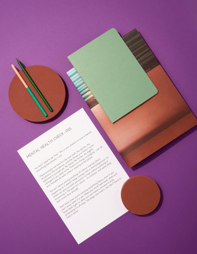 Op de afbeelding: Een stilleven op een paarse ondergrond met een groen notitieboek, een bruin notitieboek, een vel papier met de tekst "MENTAL HEALTH CHECK-INS", twee potloden en twee bruine onderzetters. De potloden zijn groen en oranje.