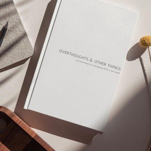 Puede incluir: Un diario blanco con el texto "OVERTHOUGHTS & OTHER THINGS" y "journal prompts for untangling what's in your head" se muestra. Un lápiz y un pequeño bloc de notas son visibles en el fondo. Una bandeja de madera y una flor seca también están en la imagen.