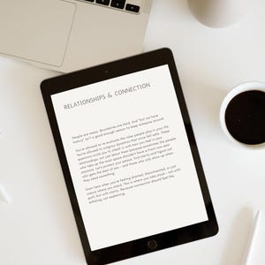 Op de afbeelding: Een zwarte tablet met tekst over relaties en verbinding, samen met een laptop, een koffiekopje en een decoratieve vaas op een wit oppervlak. De tekst op de tablet luidt "RELATIONSHIPS & CONNECTION". Een pen is ook zichtbaar.