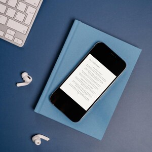 Op de afbeelding: Een bovenaanzicht van een werkruimte met een blauwe achtergrond. Een zilverkleurig toetsenbord staat linksboven. Een blauw boek en een smartphone met de tekst "SELF-DISCOVERY" staan in het midden. Twee witte draadloze oordopjes liggen verspreid.