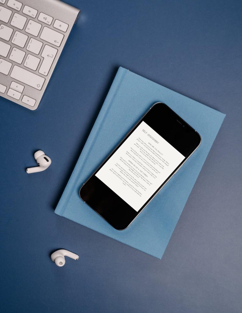 Op de afbeelding: Een bovenaanzicht van een werkruimte met een blauwe achtergrond. Een zilverkleurig toetsenbord staat linksboven. Een blauw boek en een smartphone met de tekst "SELF-DISCOVERY" staan in het midden. Twee witte draadloze oordopjes liggen verspreid.