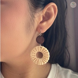 Puede incluir: Pendientes colgantes con un disco de madera marrón oscuro sobre un diseño circular beige tejido. Los pendientes cuelgan de ganchos plateados. Los pendientes son de estilo natural y bohemio.