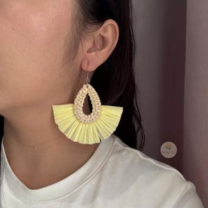Puede incluir: Pendientes de rafia amarillos con un diseño de lágrima tejido. Los pendientes tienen forma de abanico con un color amarillo claro y un centro tejido beige natural. Los pendientes están suspendidos de ganchos plateados.