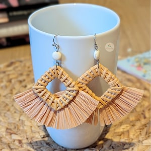 Puede incluir: Un par de pendientes hechos a mano con un diseño tejido natural. Los pendientes presentan un centro cuadrado tejido con una franja de rafia de color melocotón claro. Los pendientes cuelgan de una taza blanca.