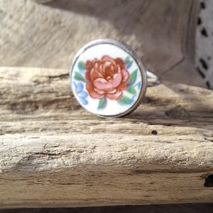 Könnte beinhalten: Silberring mit rundem, weißem Gesicht mit einer handgemalten roten Rose mit grünen Blättern und blauen Akzenten. Eine passende Anhänger-Halskette ist im Hintergrund zu sehen. Der Schmuck ist auf einer rustikalen Holzoberfläche platziert.