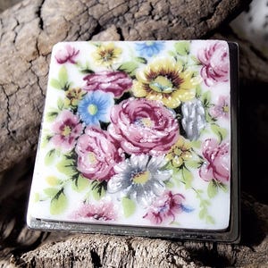 Pode incluir: Um azulejo quadrado com moldura prateada com um design floral. O azulejo apresenta um fundo branco com flores rosa, amarelas e azuis. As flores são cercadas por folhas verdes.