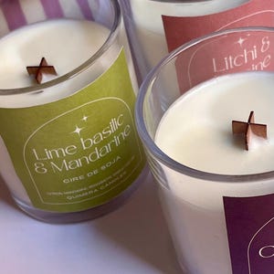 May include: Three soy wax candles in clear glass jars with wooden wicks. The labels are green, pink, and purple, with text that reads "Lime Basilic & Mandarine", "Litchi & ine", and "C". The candles have a creamy white wax.