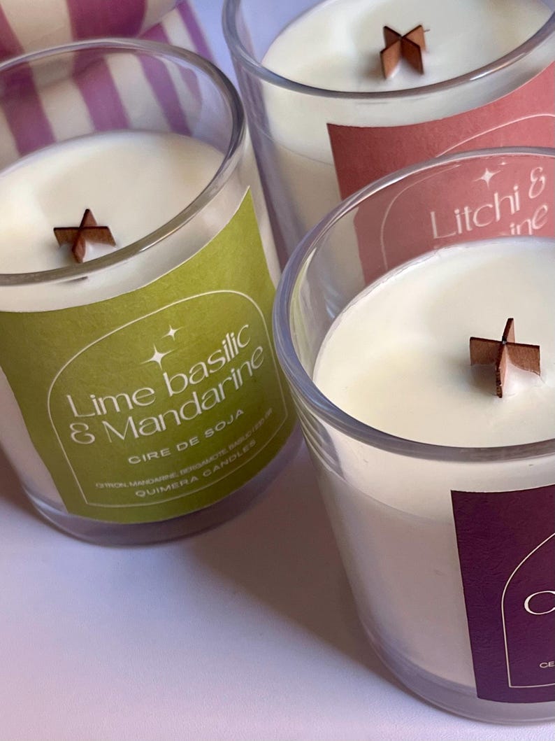 May include: Three soy wax candles in clear glass jars with wooden wicks. The labels are green, pink, and purple, with text that reads "Lime Basilic & Mandarine", "Litchi & ine", and "C". The candles have a creamy white wax.