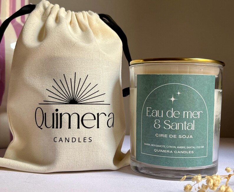 May include: A candle in a glass jar with a gold rim, labelled "Eau de mer & Santal" and "Cire de Soja." Beside it is a cream drawstring bag with the "Quimera Candles" logo. The candle is a soy wax candle.