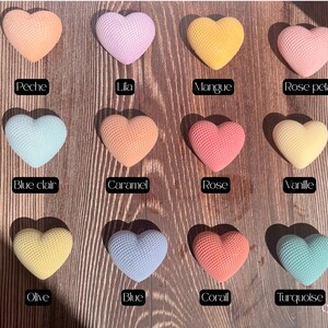 May include: A collection of heart-shaped objects in various colors, including peach, lilac, yellow, and pink. Each heart has a textured surface and is labeled with a color name. The objects are arranged on a wooden surface.