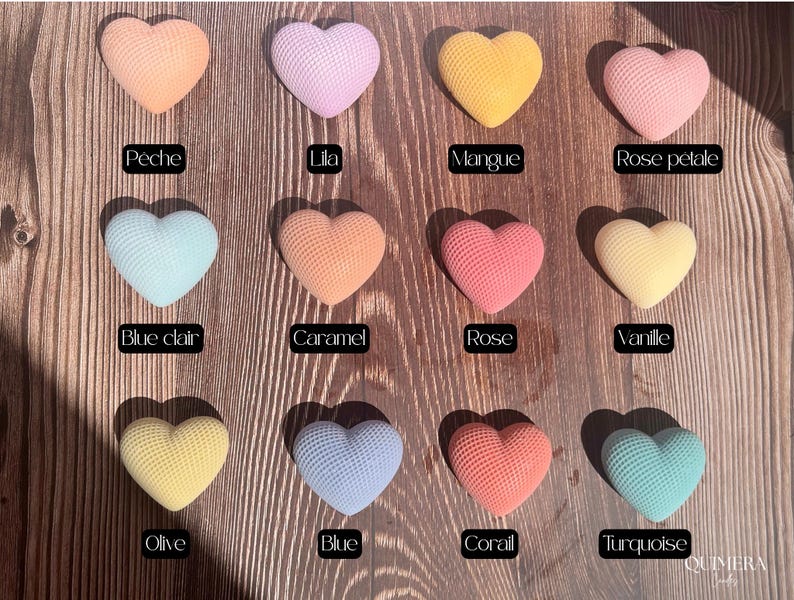 May include: A collection of heart-shaped objects in various colors, including peach, lilac, yellow, and pink. Each heart has a textured surface and is labeled with a color name. The objects are arranged on a wooden surface.