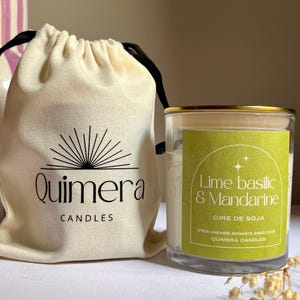 May include: A soy wax candle in a glass jar with a gold rim, next to a cream-colored drawstring bag. The candle has a green label with the text "Lime Basilic & Mandarine". The bag is printed with the brand name "Quimera Candles".