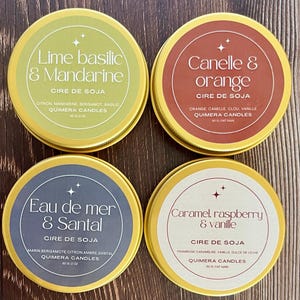 May include: Four round, gold-rimmed soy wax candles in tins. The labels feature text in French, including "Lime basilic & Mandarine," "Canelle & orange," "Eau de mer & Santal," and "Caramel, raspberry & vanille." Each tin is 60g / 2 oz.