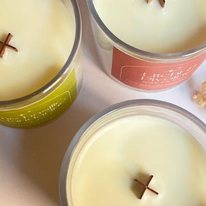 May include: Three clear glass candles with creamy wax and wooden cross-shaped wicks. One candle has a green label, another has a red label, and the third has no visible label. The candles are likely scented and designed for home fragrance.