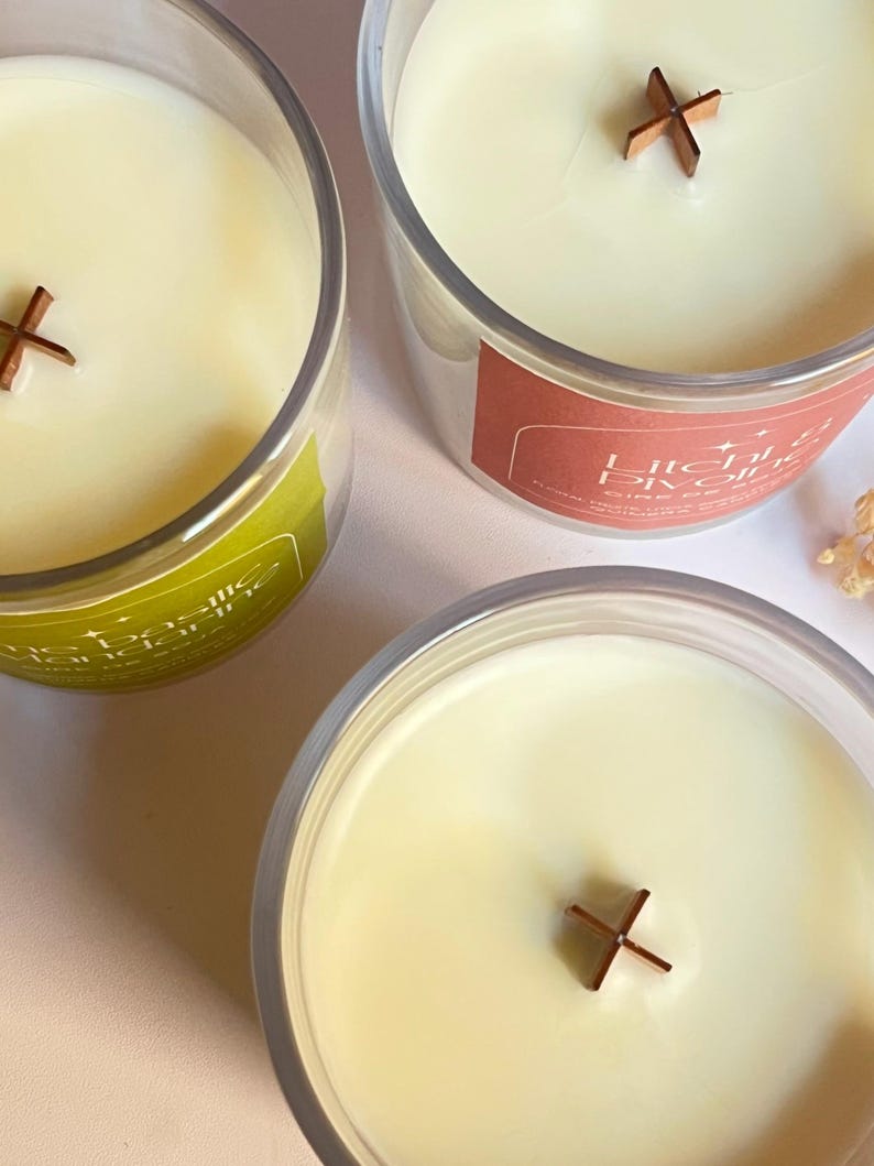 May include: Three clear glass candles with creamy wax and wooden cross-shaped wicks. One candle has a green label, another has a red label, and the third has no visible label. The candles are likely scented and designed for home fragrance.
