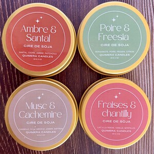 May include: Four round, gold-rimmed candle tins with labels. The labels are in French and feature the scents: Ambre & Santal, Poire & Freesia, Musc & Cachemire, and Fraises & chantilly. Each tin contains 2 oz of soy wax.