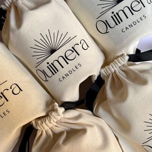 May include: Close-up of several off-white drawstring bags with black text and a sunburst design. The bags are labeled "Quimera Candles". The bags have black drawstrings.