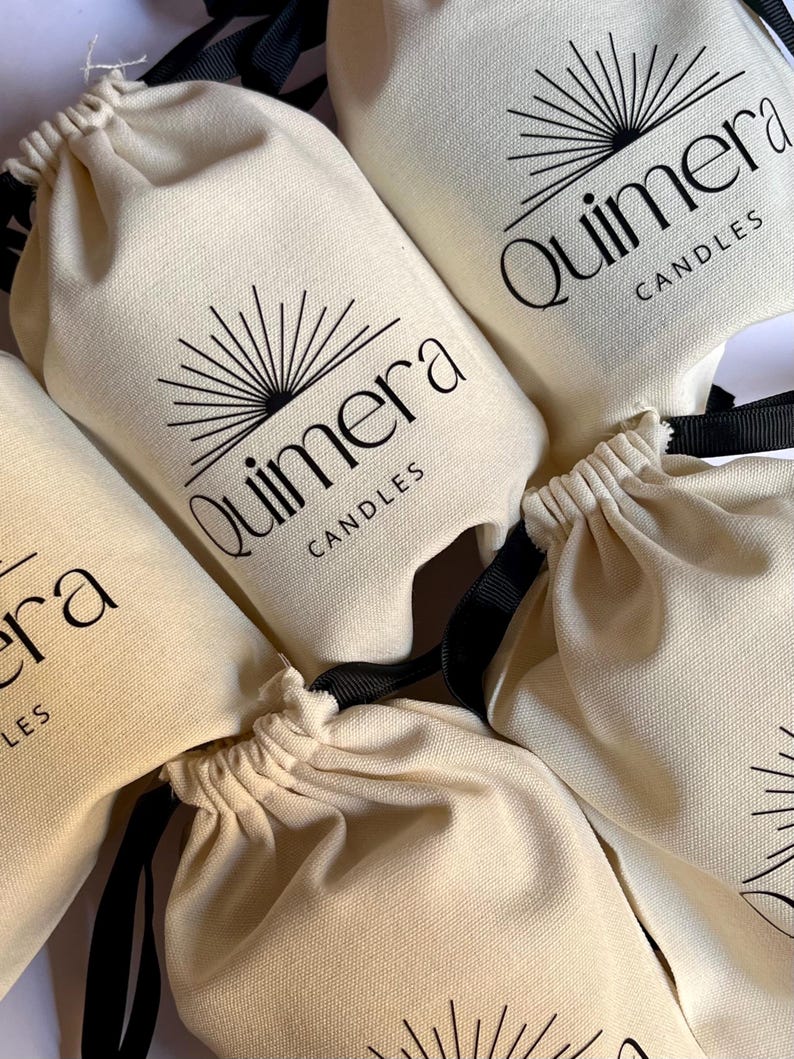 May include: Close-up of several off-white drawstring bags with black text and a sunburst design. The bags are labeled "Quimera Candles". The bags have black drawstrings.