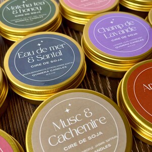 May include: A collection of soy wax candles in gold tins with various scents. Each candle has a label with the scent name, such as "Matcha Tea & Honey" and "Eau de Mer & Santal". The labels are in different colors, including green, purple, and brown.