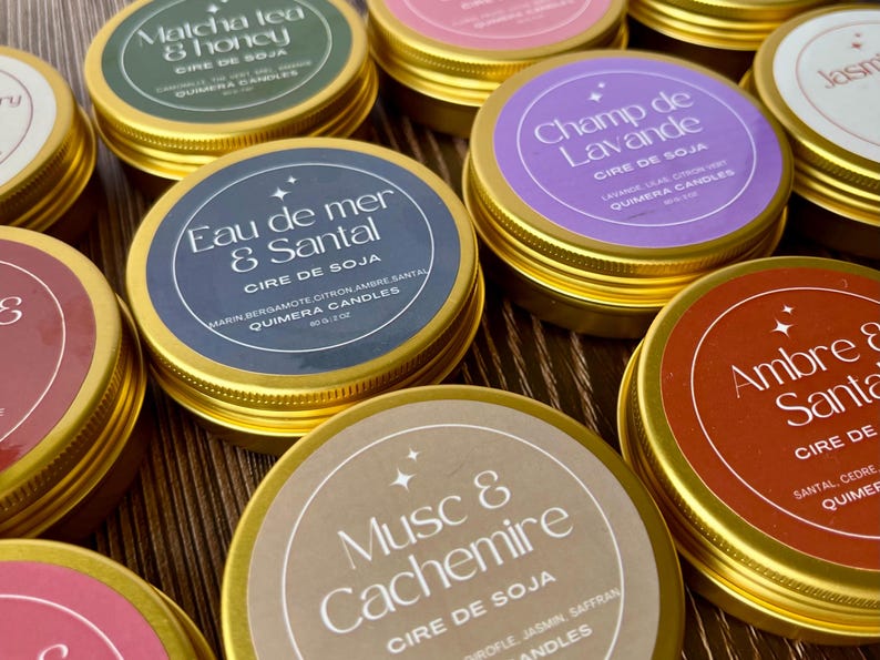 May include: A collection of soy wax candles in gold tins with various scents. Each candle has a label with the scent name, such as "Matcha Tea & Honey" and "Eau de Mer & Santal". The labels are in different colors, including green, purple, and brown.