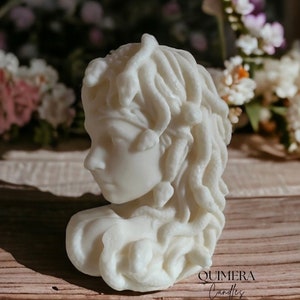 Könnte beinhalten: Eine weiße Kerze in Form einer Büste der Medusa, mit detaillierten Locken und Gesichtszügen. Die Kerze steht vor einem verschwommenen Hintergrund aus Blumen und einer Holzoberfläche. Der Text "QUIMERA Candles" ist unten sichtbar.