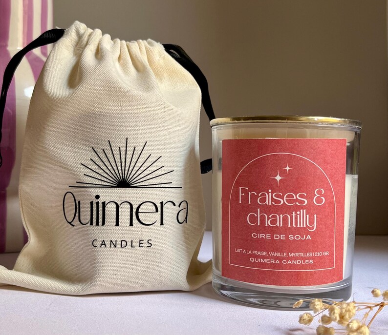 May include: A candle in a clear glass jar with a gold rim, labelled "Fraises & chantilly" with a red label. Beside it is a beige drawstring bag with the "Quimera Candles" logo.