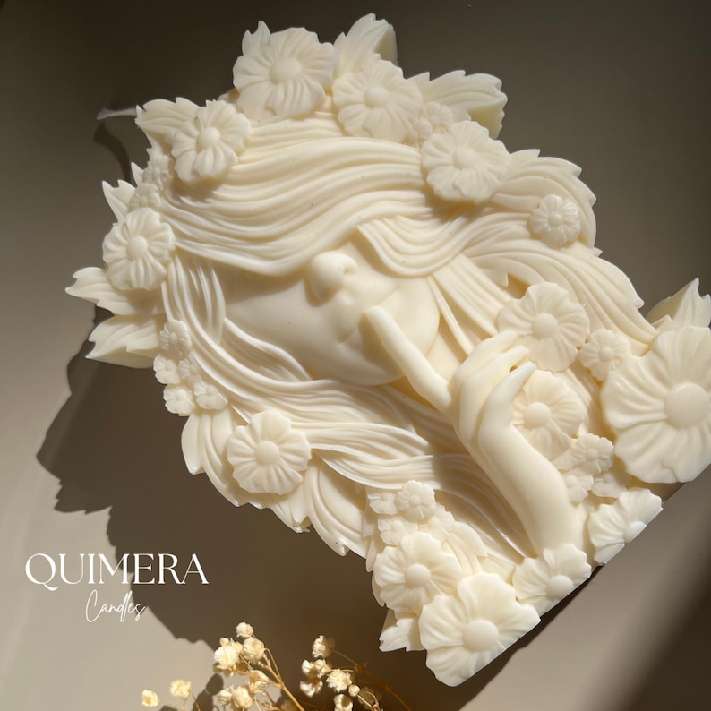May include: Ivory-colored candle shaped like a woman's face with flowing hair and floral accents. The candle features the text "QUIMERA Candles" in a script font. The woman's face is in profile, with a finger touching her lips.