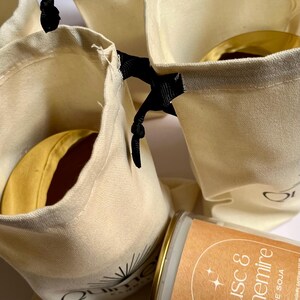 May include: Several small, cream-colored drawstring bags with black ribbon closures. Each bag contains a round, yellow item with a brown center. A candle with a brown label that reads "Musc & Cachemire" is also visible.