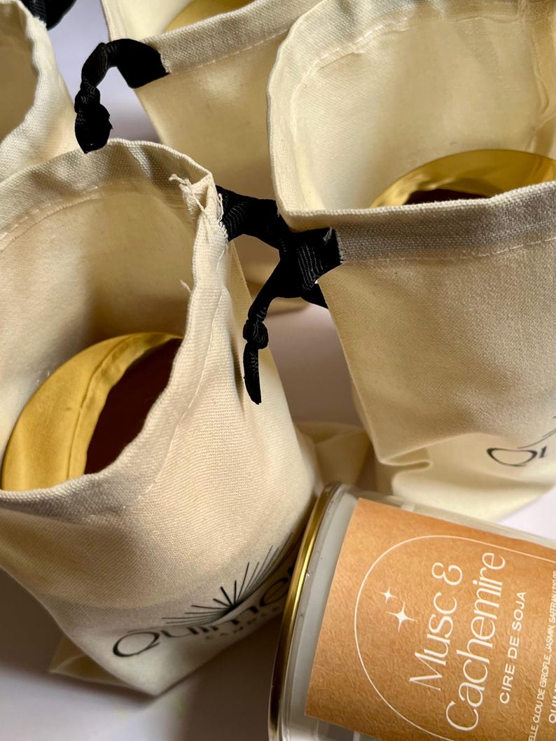 May include: Several small, cream-colored drawstring bags with black ribbon closures. Each bag contains a round, yellow item with a brown center. A candle with a brown label that reads "Musc & Cachemire" is also visible.