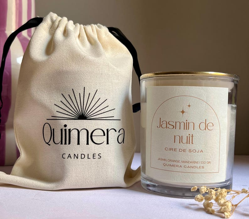 Pode incluir: Um frasco de vela de vidro transparente com uma borda dourada e um r&oacute;tulo que diz "Jasmin de nuit". Ao lado, uma bolsa com cord&atilde;o creme com o logotipo "Quimera Candles". A vela &eacute; preenchida com cera e a bolsa &eacute; de lona.