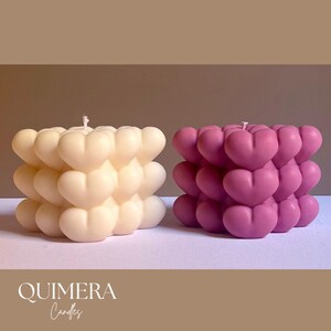 May include: Two heart-shaped candles. One is ivory, the other is a deep pink. Each candle is composed of multiple heart shapes stacked together. The candles are on a white surface with the text "QUIMERA Candles" at the bottom.