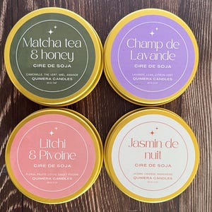 May include: Four round, gold-rimmed soy wax candles with labels. The labels feature text in various colors, including green, purple, pink, and white, with the scents "Matcha tea & honey", "Champ de Lavande", "Litchi & Pivoine", and "Jasmin de nuit".