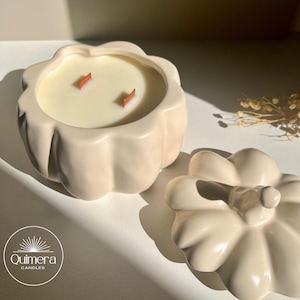 K&ouml;nnte beinhalten: Eine beige k&uuml;rbisf&ouml;rmige Kerze mit zwei Holzdochten. Die Kerze befindet sich in einem passenden Keramikbeh&auml;lter, mit einem Deckel in der N&auml;he. Die Kerze ist von Quimera Candles.