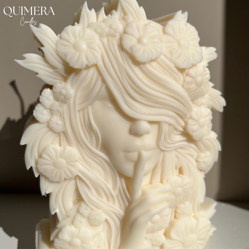 May include: Ivory-colored candle shaped like a woman's face with a finger to her lips, surrounded by floral details. The candle has the text "QUIMERA Candles" in the upper left corner.