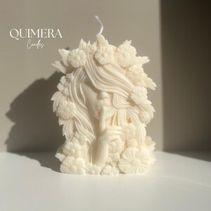May include: Ivory-colored candle shaped like a woman with long flowing hair and floral accents. The woman is depicted with a finger pressed to her lips, in a gesture of silence. The candle has the text "QUIMERA Candles" on the upper left.