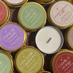 May include: Overhead shot of several small, round candles in gold tins. The labels feature various scents like "Lime Basilic & Mandarine" and "Caramel, raspberry & vanille". One candle is lit, showing a wooden wick.