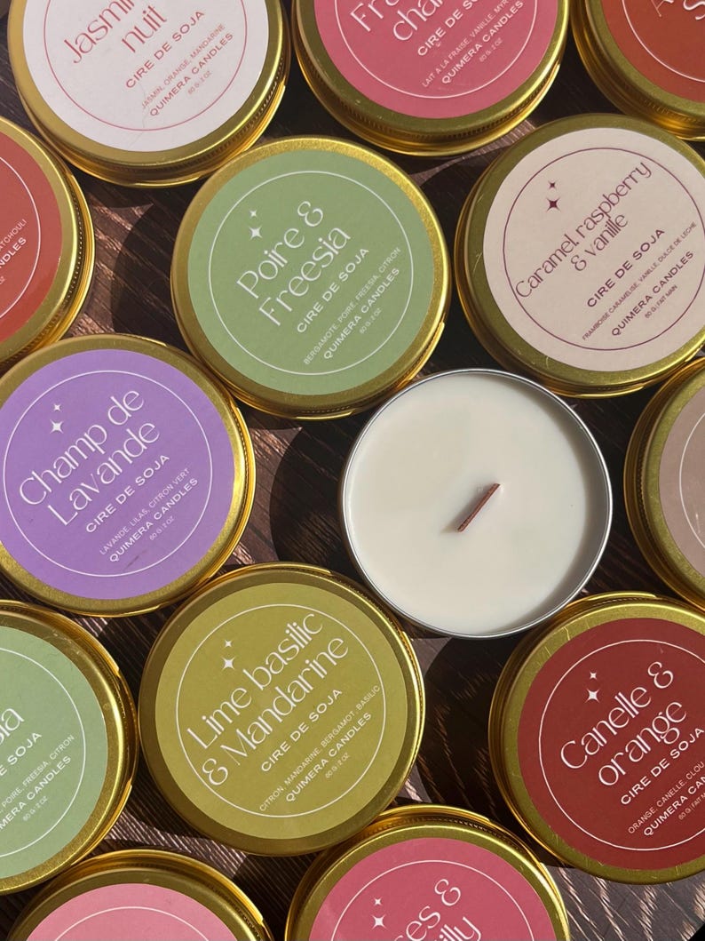 May include: Overhead shot of several small, round candles in gold tins. The labels feature various scents like "Lime Basilic & Mandarine" and "Caramel, raspberry & vanille". One candle is lit, showing a wooden wick.