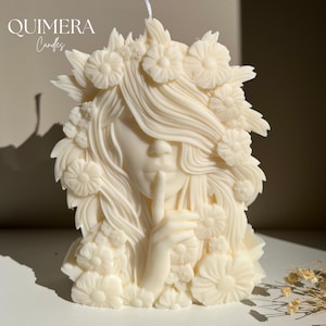 May include: Ivory-colored candle shaped like a woman with a finger to her lips, surrounded by floral details. The candle has long flowing hair and is adorned with flower accents. The text "QUIMERA Candles" is at the top left.
