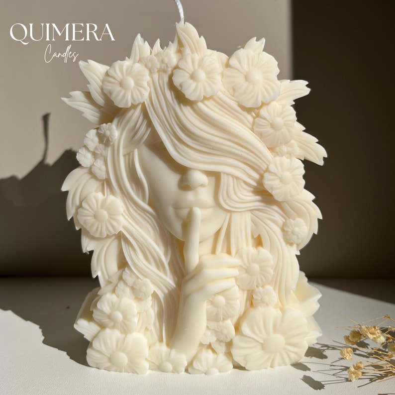 May include: Ivory-colored candle shaped like a woman with a finger to her lips, surrounded by floral details. The candle has long flowing hair and is adorned with flower accents. The text "QUIMERA Candles" is at the top left.