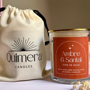 May include: A clear glass candle jar with a gold rim and a red label that reads "Ambre & Santal". The candle is next to a cream-colored drawstring bag with the "Quimera Candles" logo. The candle is made with soy wax.